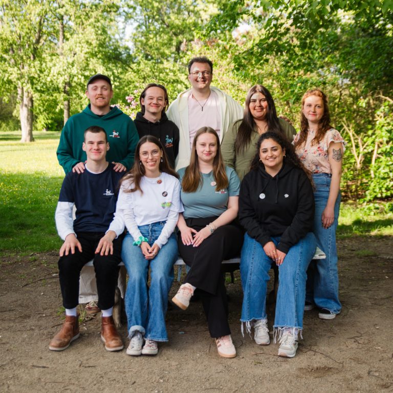 Gruppenfoto von neun Personen in einem grünen Park. Vier Personen sitzen auf einer Bank im Vordergrund, fünf stehen dahinter.